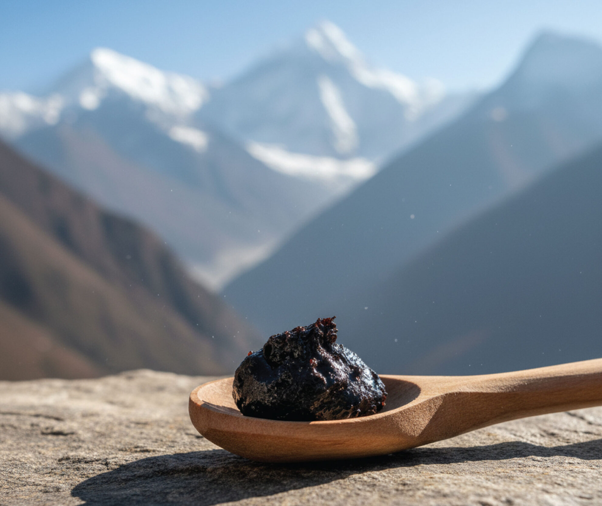 A wooden spoon holding a dark paste, possibly Shilajit, rests on a rock with blurred snow-capped mountains and a clear blue sky in the background.
