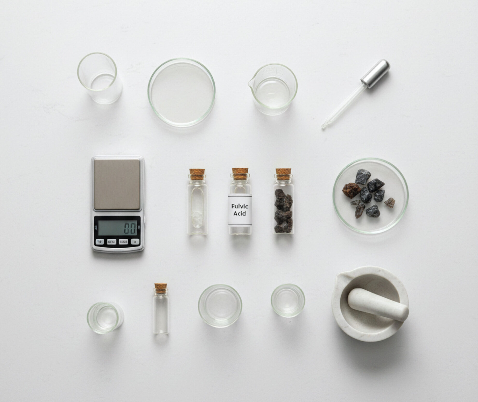 A flat lay of laboratory glassware, a digital scale, a mortar and pestle, a dropper, and three labeled vials (one marked “Fulvic Acid”) with solids inside, all arranged neatly on a white surface.