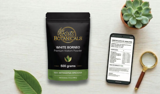 A package of Kats Botanicals White Borneo Kratom powder sits on a table next to a green leaf, a magnifying glass, a succulent plant, and a smartphone displaying a certificate of analysis.
