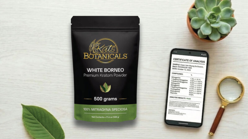 A package of Kats Botanicals White Borneo Kratom powder sits on a table next to a green leaf, a magnifying glass, a succulent plant, and a smartphone displaying a certificate of analysis.