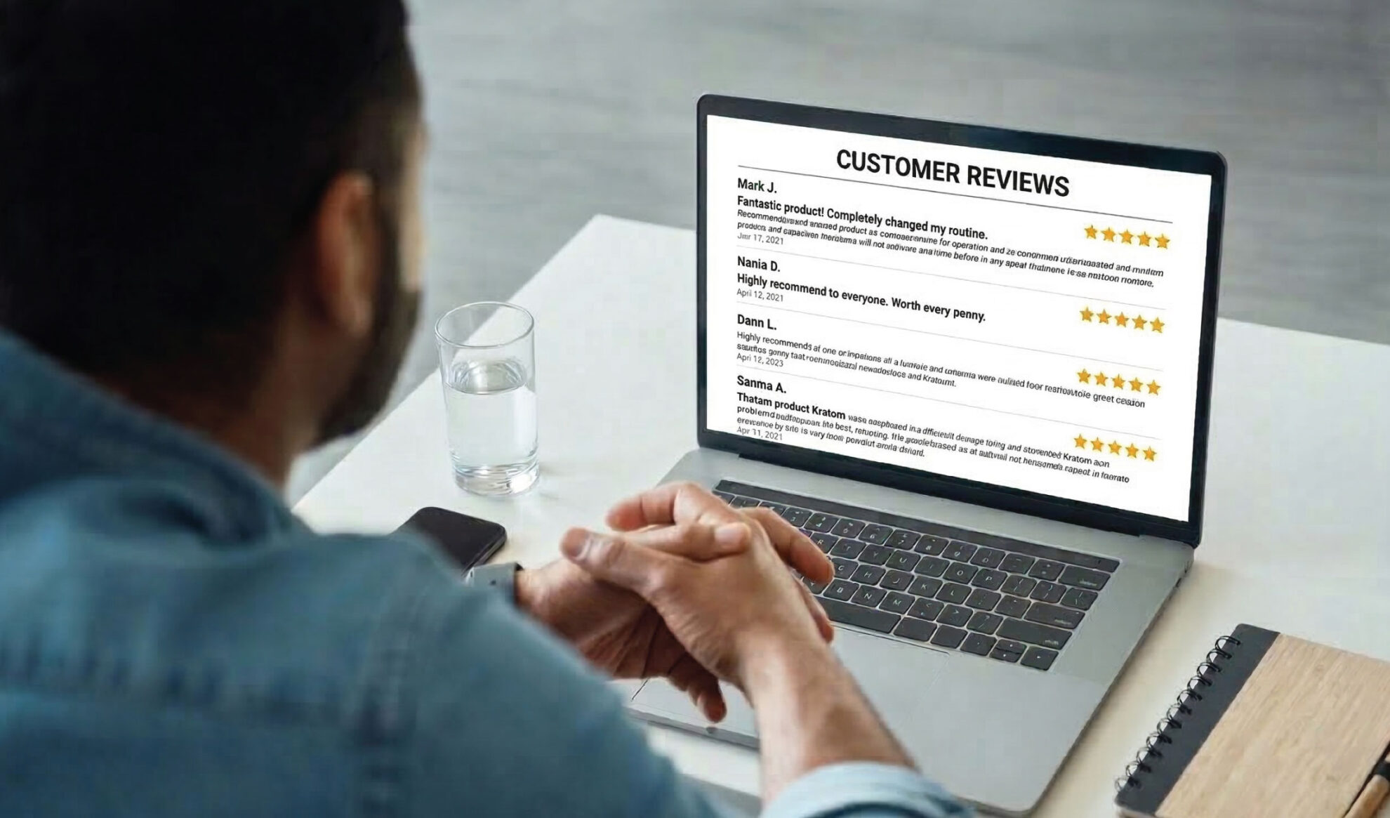 A person sitting at a desk with a laptop open, reading customer reviews displayed on the screen. A notebook, pen, and glass of water are also on the desk. The reviews are rated with five stars.