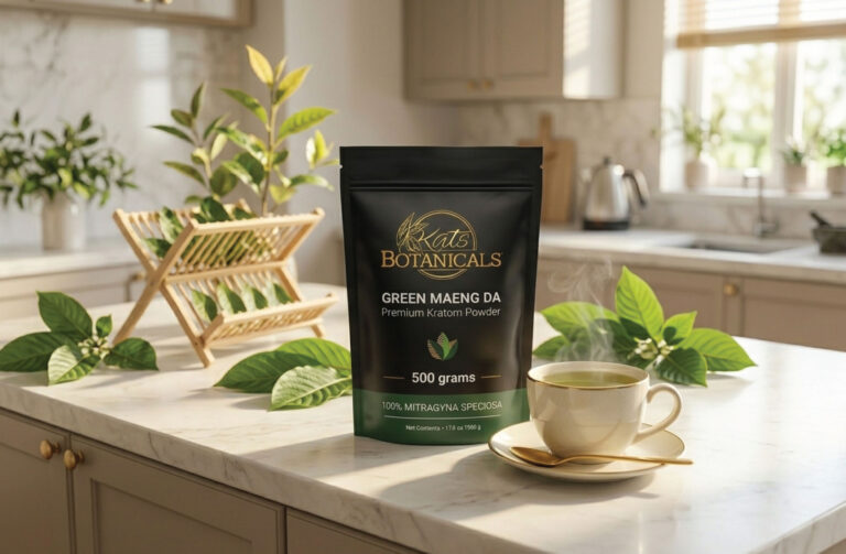 A bag of Kat’s Botanicals Green Maeng Da kratom powder sits on a marble kitchen counter next to a steaming cup, saucer, and spoon, with green leaves and kitchen items in the background.
