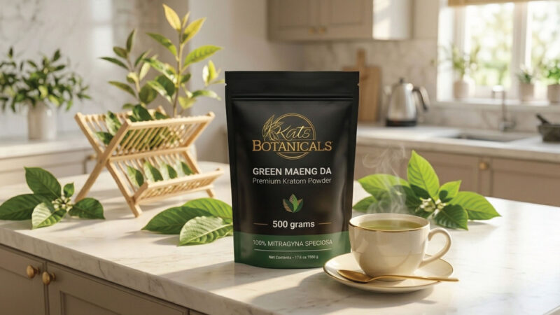 A bag of Kat’s Botanicals Green Maeng Da kratom powder sits on a marble kitchen counter next to a steaming cup, saucer, and spoon, with green leaves and kitchen items in the background.