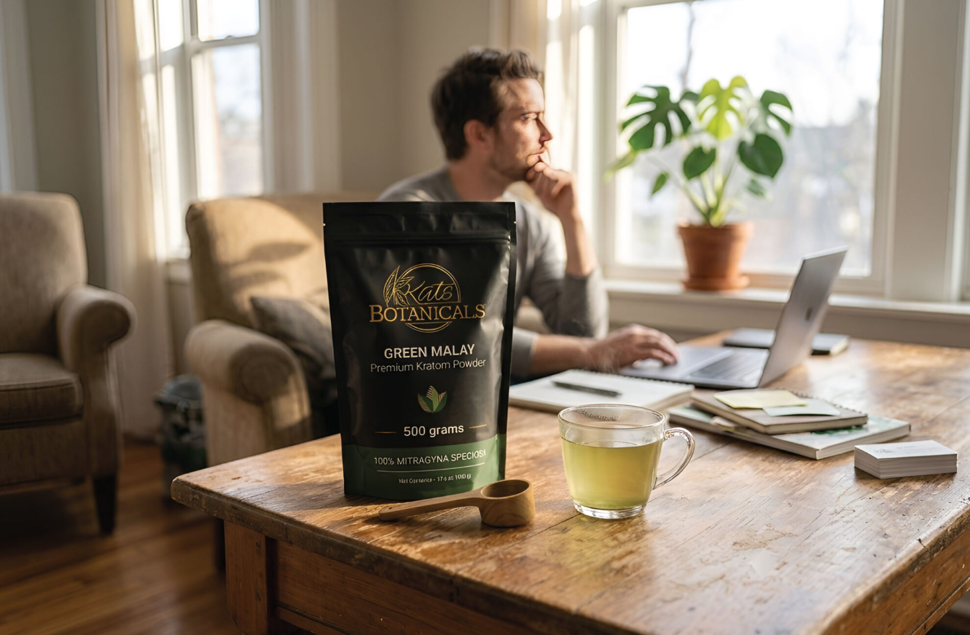 A man sits at a desk working on a laptop, with a cup of green tea and a bag of Kat’s Botanicals Green Malay Kratom powder on a wooden table in a bright room.