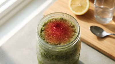 A jar of green smoothie with a red center sits on a light counter next to a half lemon, a spoon, a clear glass, and a wooden cutting board. Sunlight streams in from the left.