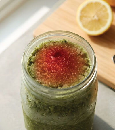 A jar of green smoothie with a red center sits on a light counter next to a half lemon, a spoon, a clear glass, and a wooden cutting board. Sunlight streams in from the left.
