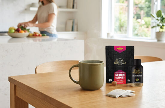 A steaming mug, kratom gummies, and a kratom shot sit on a wooden table. In the background, a woman prepares food in a bright, modern kitchen.
