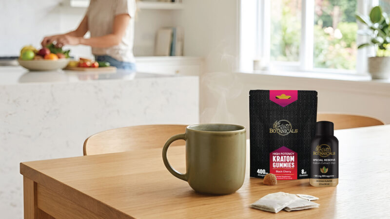 A steaming mug, kratom gummies, and a kratom shot sit on a wooden table. In the background, a woman prepares food in a bright, modern kitchen.