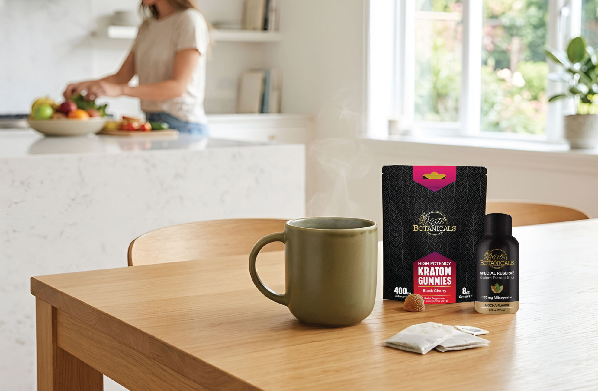 A steaming mug, kratom gummies, and a kratom shot sit on a wooden table. In the background, a woman prepares food in a bright, modern kitchen.