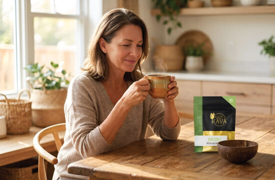 A woman sits at a wooden table in a cozy kitchen, smiling and holding a steaming mug. A bag labeled “Kava” is on the table beside her. Sunlight filters in through a window in the background.