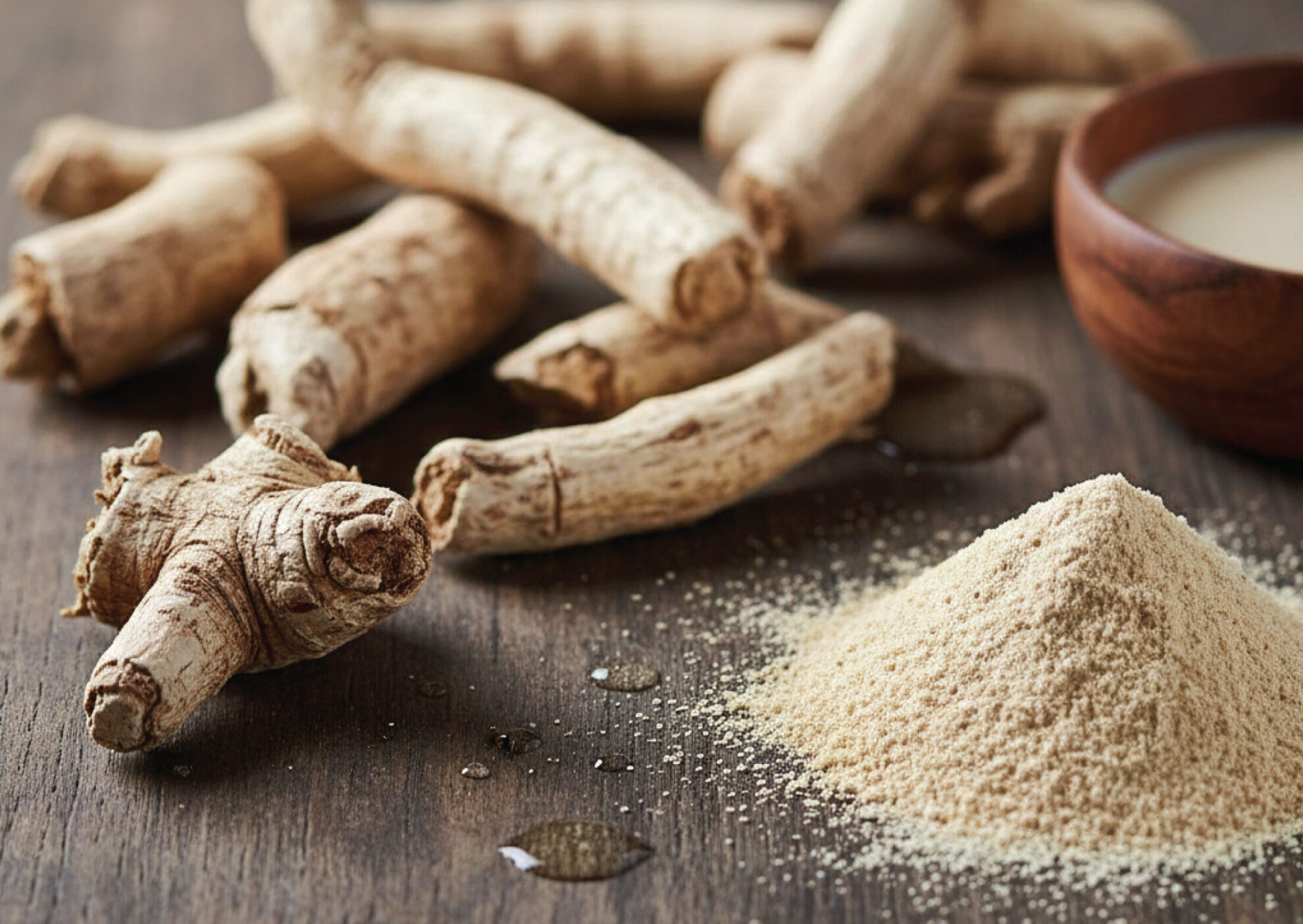 Several whole ashwagandha roots and a pile of ashwagandha powder are displayed on a wooden surface. A wooden bowl containing a creamy liquid sits in the background.