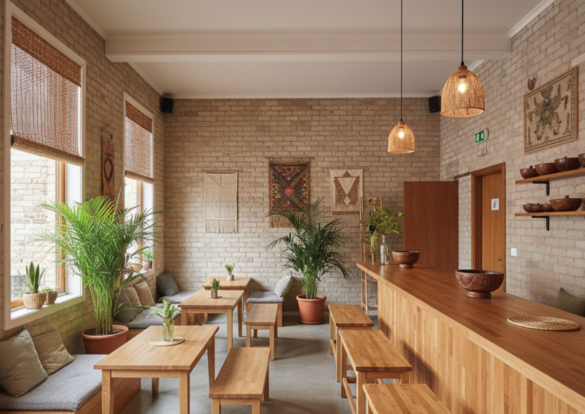 A cozy café with wooden tables and benches, potted plants, woven wall art on brick walls, and large windows with bamboo blinds. Warm pendant lights hang above the counter, creating a welcoming atmosphere.