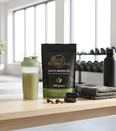 A packet of White Maeng Da kratom powder, a green shaker, towel, jump rope, earbuds, and water bottle on a wooden table in a bright gym with exercise equipment in the background.