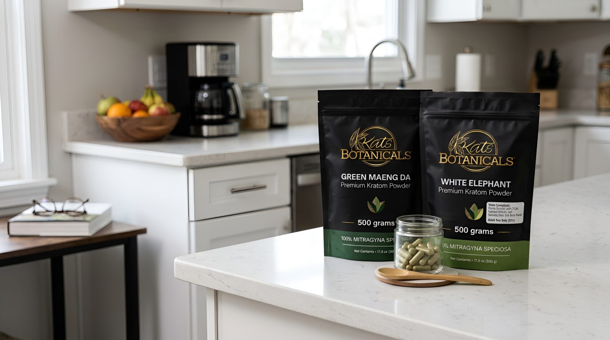 Two bags of Kats Botanicals kratom powder, Green Maeng Da and White Elephant, sit on a white kitchen counter with a small pile of capsules in front. The background shows a fruit bowl, coffee maker, and kitchen items.