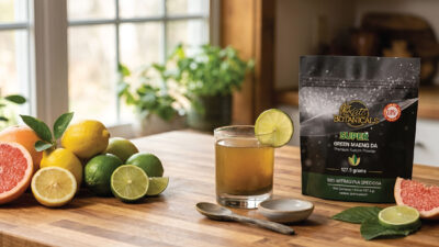 A glass of brownish drink with a lime slice sits beside citrus fruits and a packet labeled Kats Botanicals Green Maeng Da on a wooden counter in a sunlit kitchen.
