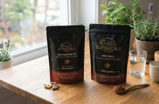 Two Kats Botanicals kratom products on a wooden table, with capsules and a spoonful of powder beside them. Potted plants and a glass of water are in the background near a sunny window.