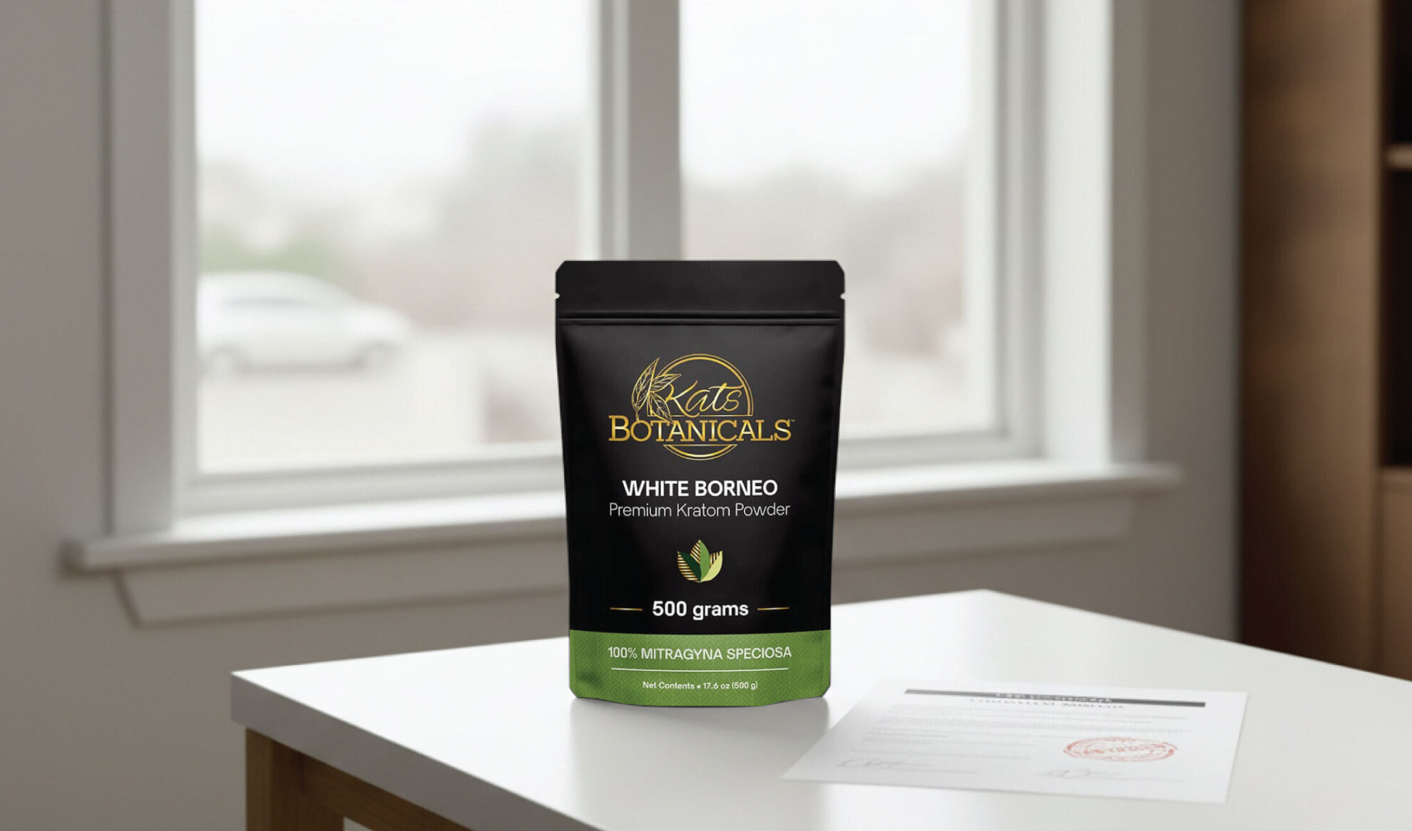 A black and green bag labeled Krats Botanicals White Borneo Premium Kratom Powder, 500 grams sits on a white table in front of a window with blurred outdoor scenery.