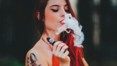 A woman with long red hair and colorful tattoos on her arm exhales vapor while holding a vape device, wearing an orange top, with a blurred dark background.