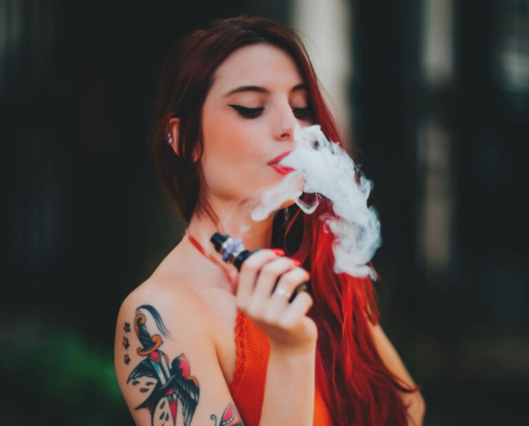 A woman with long red hair and colorful tattoos on her arm exhales vapor while holding a vape device, wearing an orange top, with a blurred dark background.