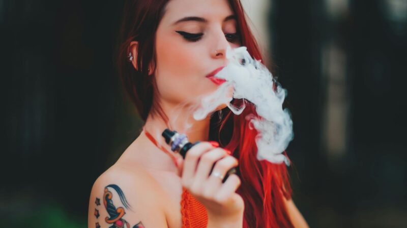 A woman with long red hair and colorful tattoos on her arm exhales vapor while holding a vape device, wearing an orange top, with a blurred dark background.