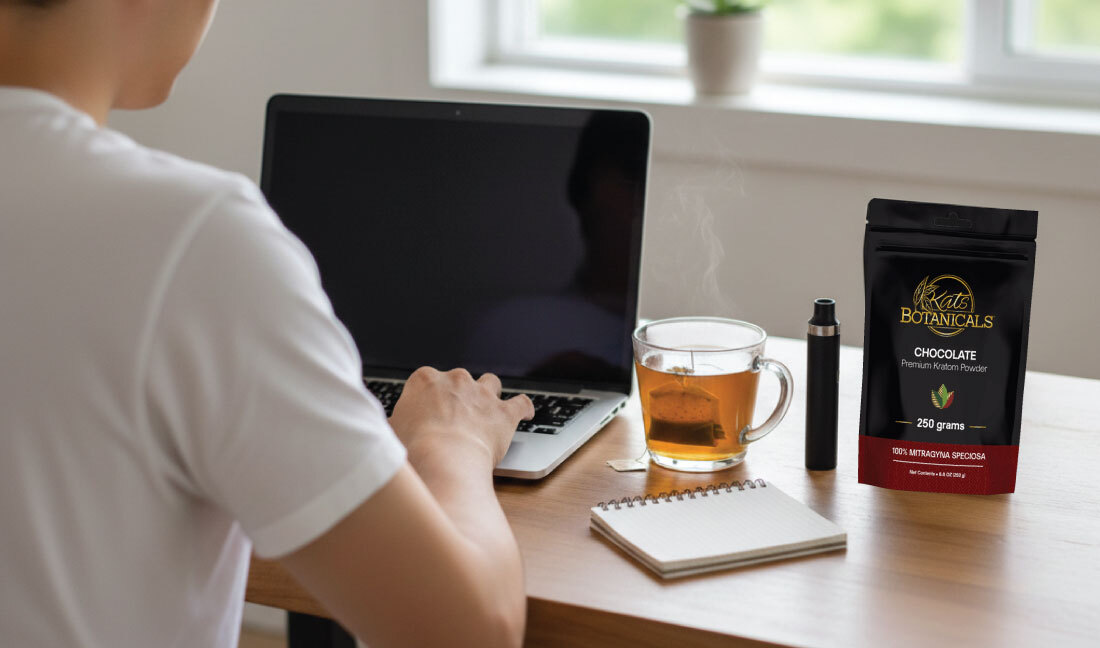 A person works on a laptop at a wooden desk with a glass of tea, a notepad, a vape pen, and a bag of Kats Botanicals chocolate powder beside them. A window in the background lets in natural light.