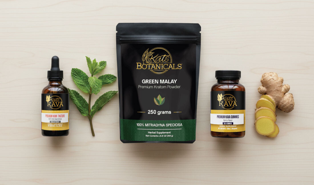 A bottle of kratom tincture, fresh mint leaves, a bag of Kats Botanicals Green Malay kratom powder, a jar of kratom gummies, and slices of ginger are arranged on a light wooden surface.