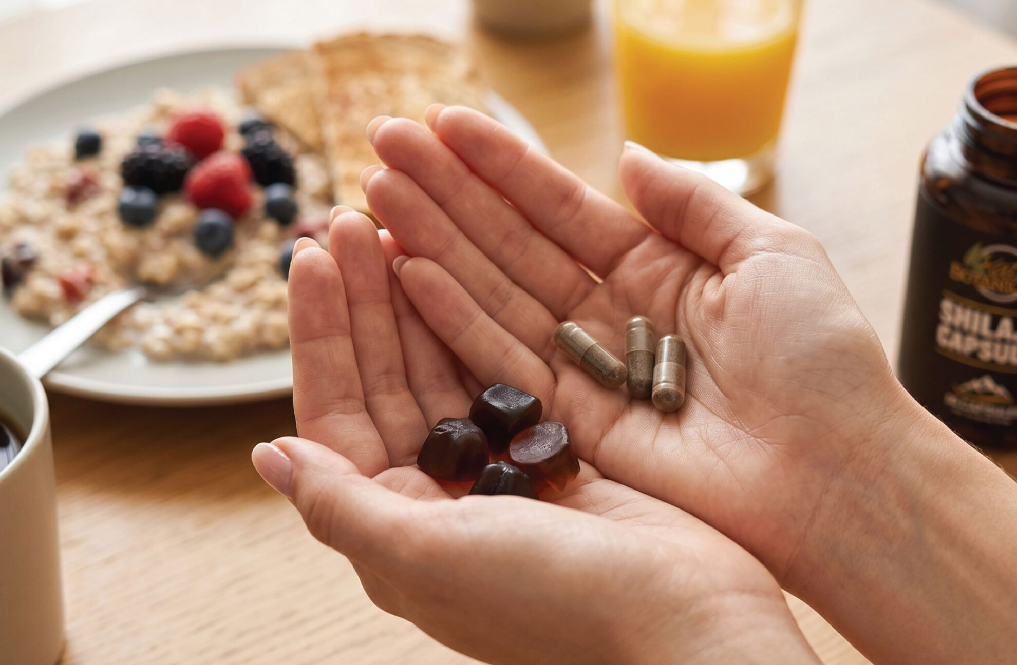 A person holds several dark gummy supplements and a few gray capsules in their hands over a wooden table set with oatmeal, fruit, a cup of coffee, and a glass of orange juice.