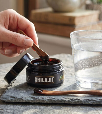 A hand scoops dark shilajit resin from a small jar labeled SHILAJIT with a wooden spoon, next to a steaming glass of water, both on a stone surface.