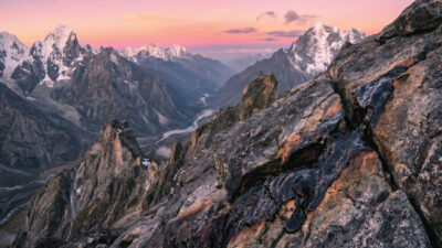 Snow-capped mountains under a colorful sunset sky, with rugged rocky terrain and valleys in the foreground. The landscape is dramatic, with patches of snow and winding river visible in the distance.