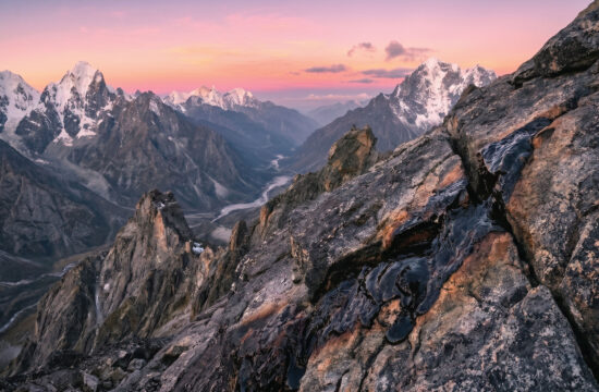 Snow-capped mountains under a colorful sunset sky, with rugged rocky terrain and valleys in the foreground. The landscape is dramatic, with patches of snow and winding river visible in the distance.