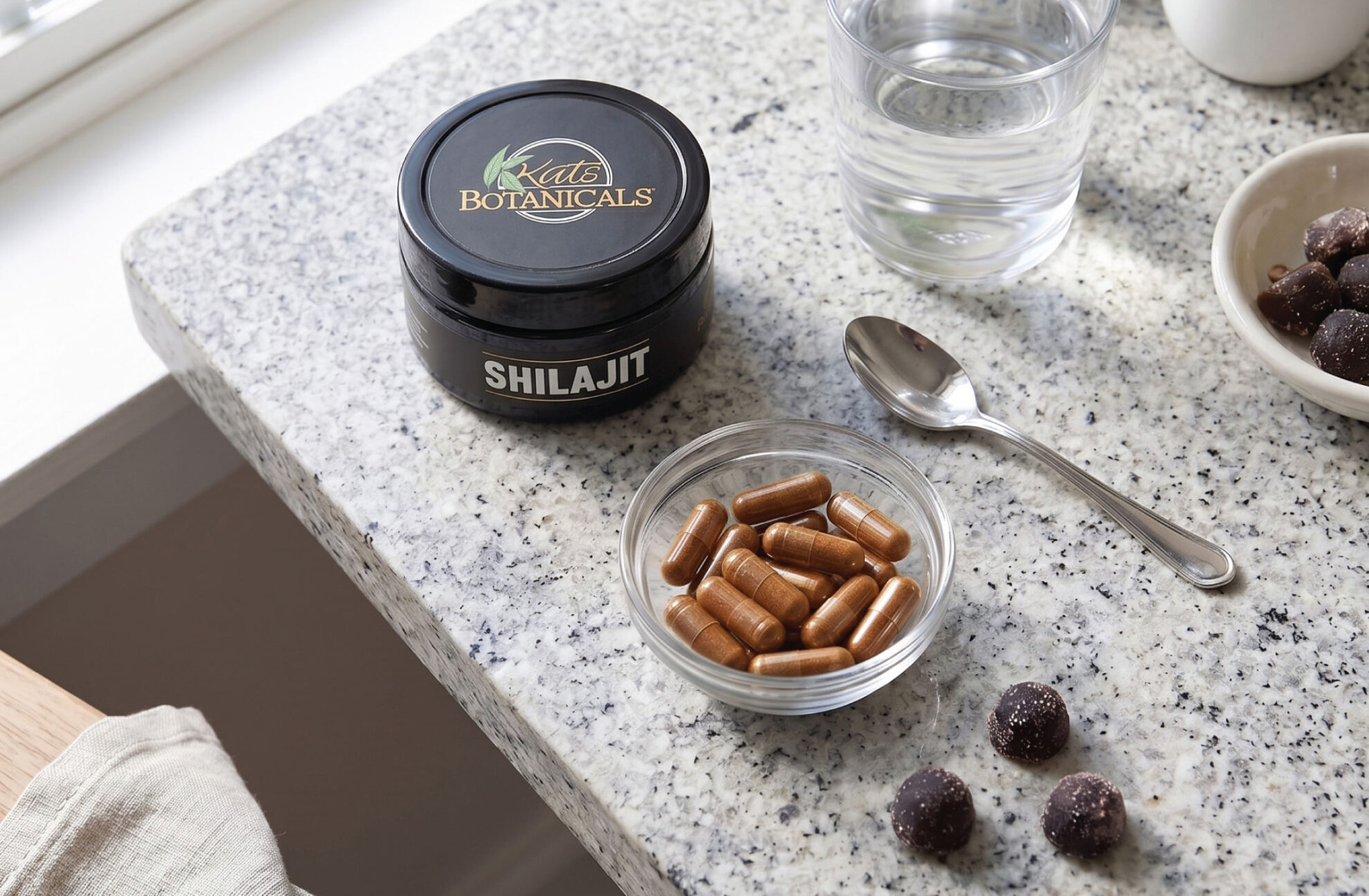 A small glass bowl of brown capsules sits on a speckled countertop next to a black jar labeled Shilajit, a spoon, a glass of water, and some dark round supplements.