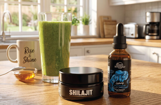 A kitchen table displays a glass of green smoothie, a mug labeled “Rise & Shine,” a dish of honey with a spoon, a black Shilajit jar, and a dropper bottle labeled “Calming” from Vigor’s Botanicals.