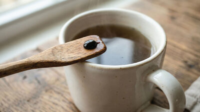 A wooden spoon with a drop of black liquid rests above a white ceramic mug filled with a dark hot beverage, placed on a rustic wooden surface. Steam rises from the cup.