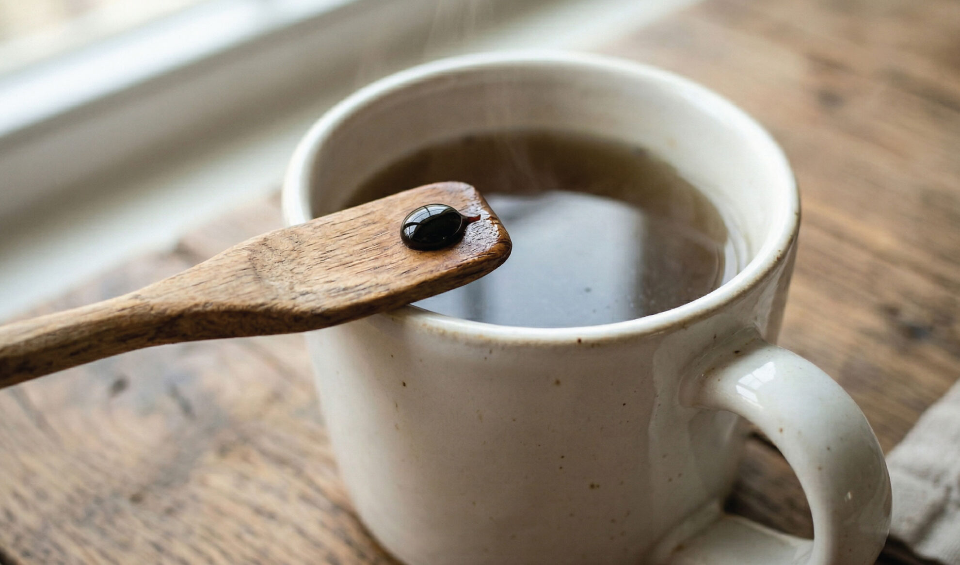 A wooden spoon with a drop of black liquid rests above a white ceramic mug filled with a dark hot beverage, placed on a rustic wooden surface. Steam rises from the cup.