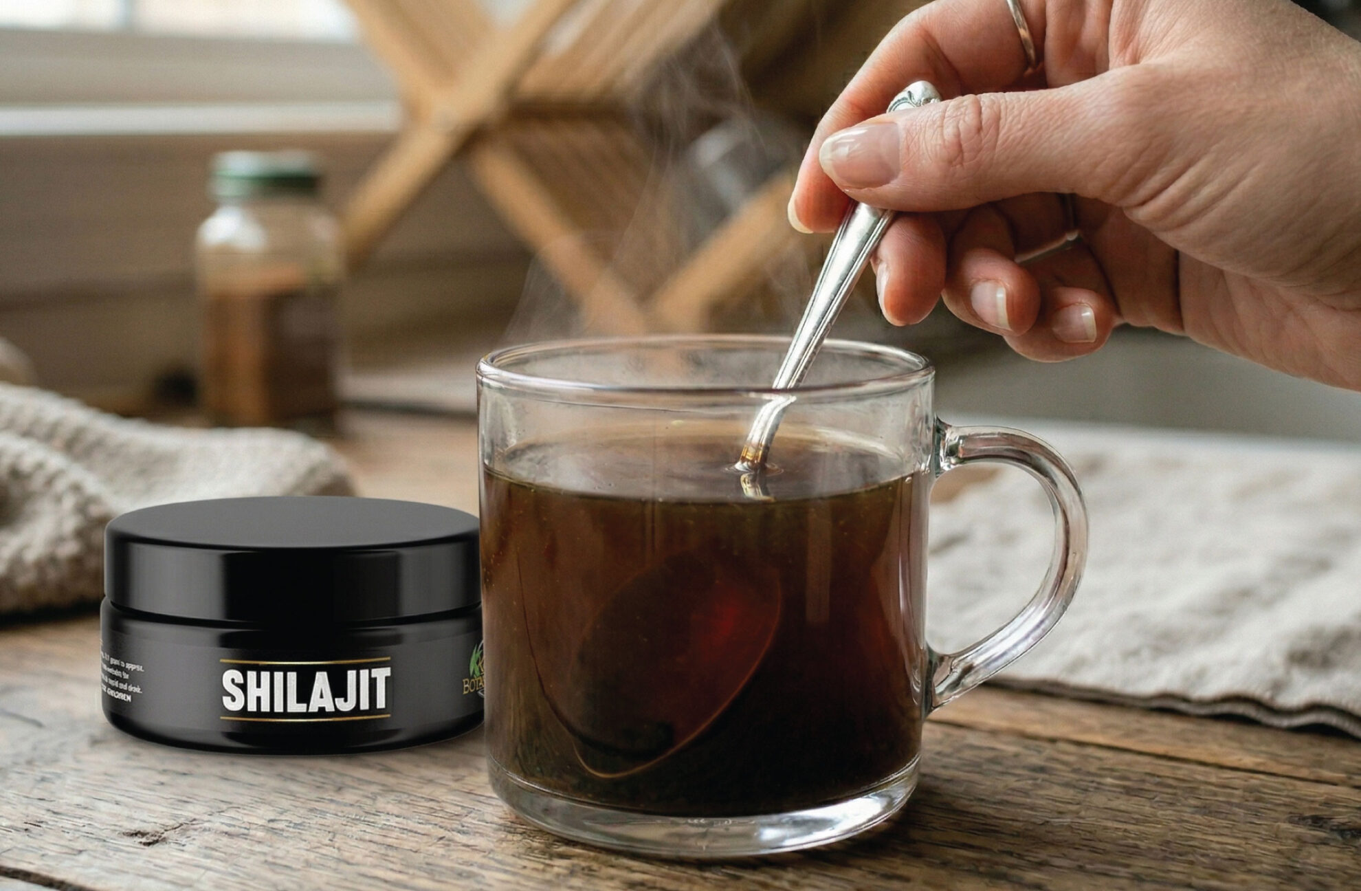  A hand stirs a steaming cup of dark tea with a spoon on a wooden table. Next to the cup is a black jar labeled Shilajit. The background shows a blurred kitchen setting.