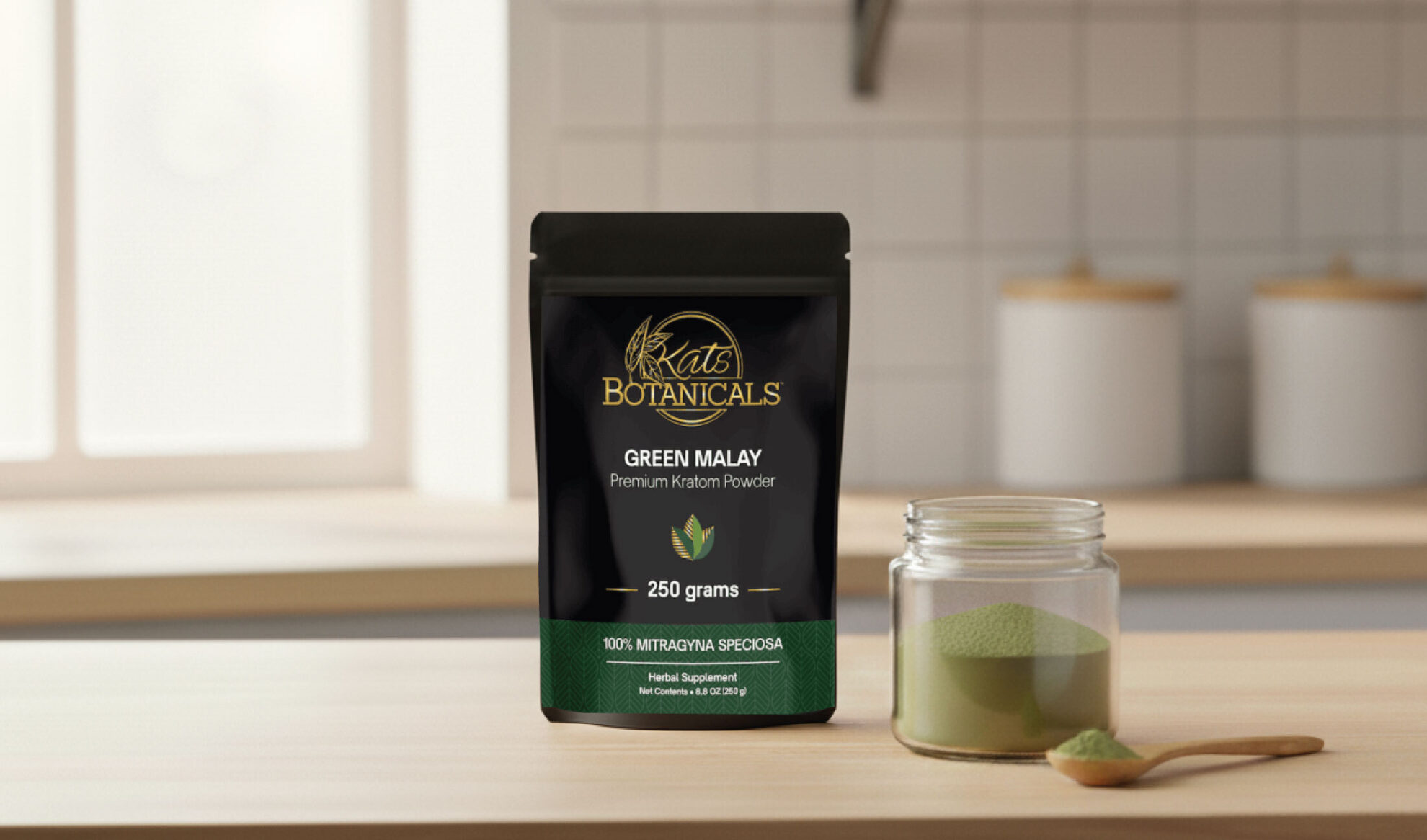 A black package of Kats Botanicals Green Malay kratom powder stands on a kitchen counter next to a glass jar and a spoon filled with green powder. Shelves and tiled walls are visible in the background.