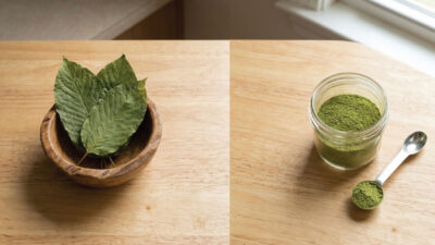 A wooden bowl with green leaves sits on a table, next to a jar filled with green powder and a metal spoon with more powder on a wooden surface by a window.
