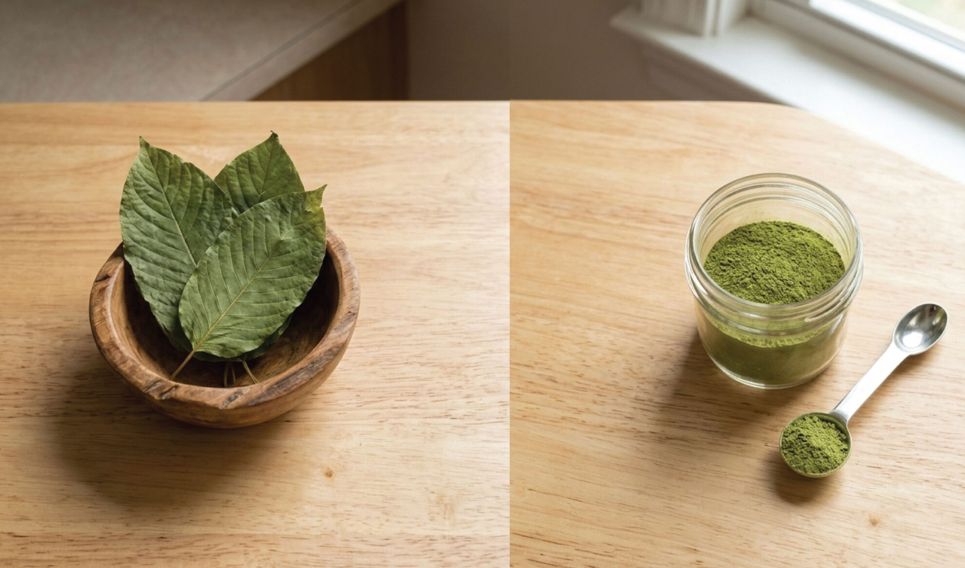 A wooden bowl with green leaves sits on a table, next to a jar filled with green powder and a metal spoon with more powder on a wooden surface by a window.