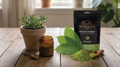 A bag of Kats Botanicals kratom powder, a glass jar, a wooden spoon, green leaves, and a potted succulent sit on a wooden table with sunlight shining through a window in the background.