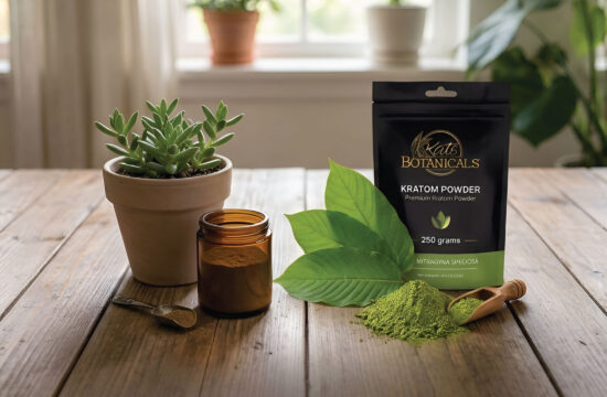 A bag of Kats Botanicals kratom powder, a glass jar, a wooden spoon, green leaves, and a potted succulent sit on a wooden table with sunlight shining through a window in the background.