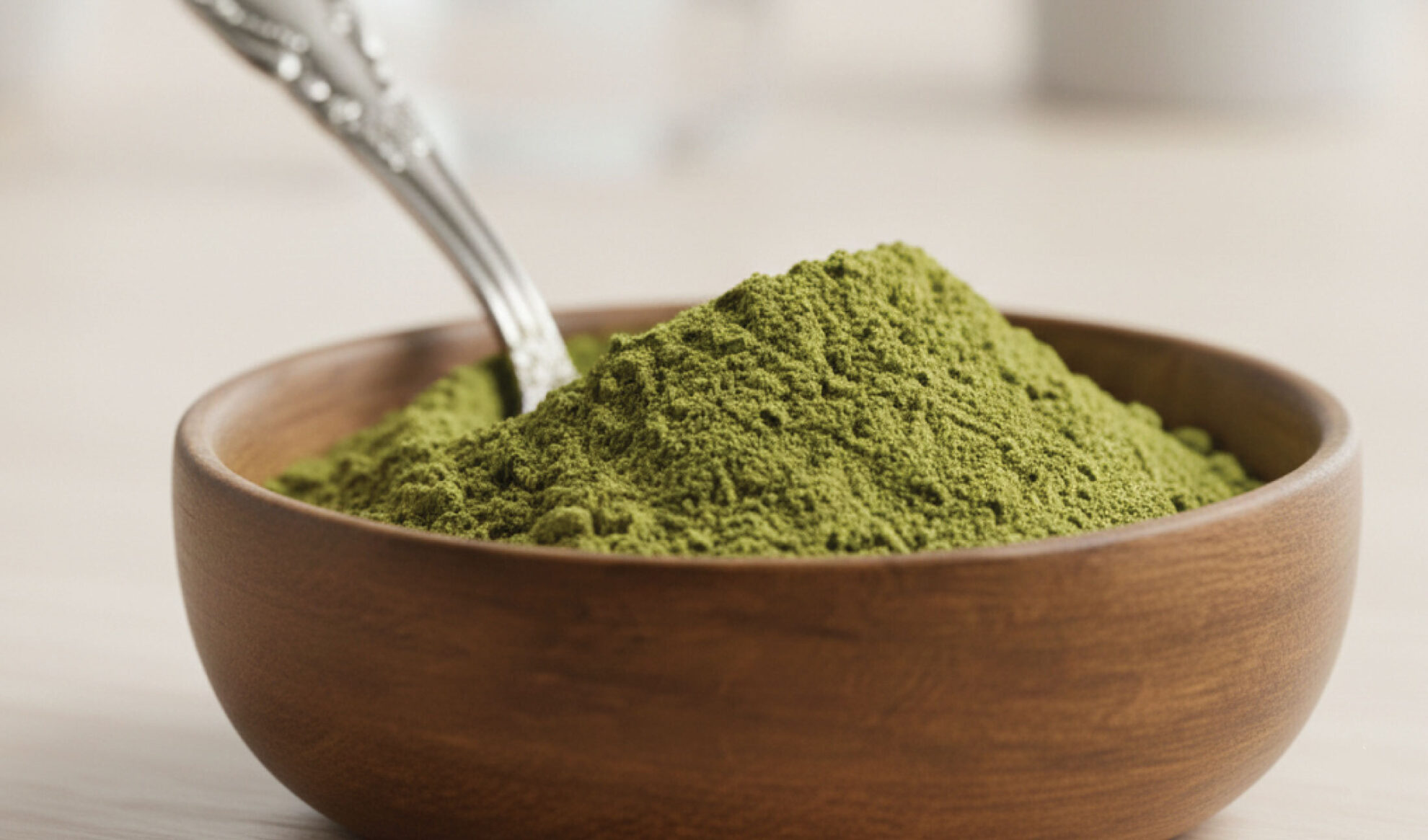 A wooden bowl filled with green powder, with a metal spoon resting inside, placed on a light-colored surface.