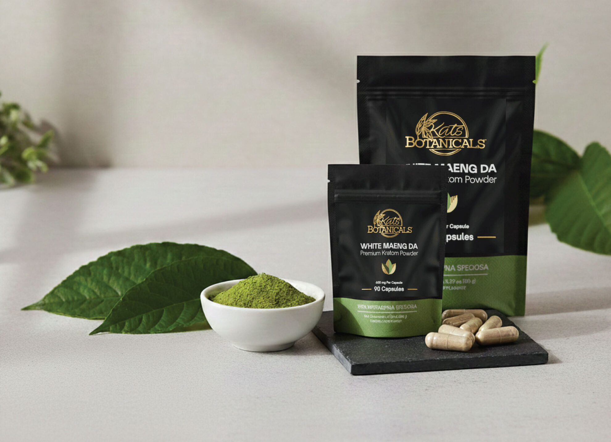 A display of Kratom Botanicals products, including two black packages, a small white bowl filled with green powder, several beige capsules, and green leaves arranged on a light surface.
