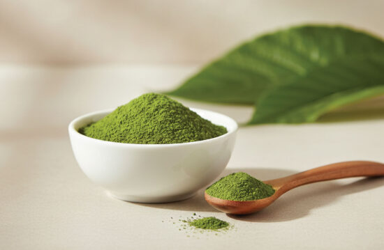 A white bowl filled with green powder sits on a light surface next to a wooden spoon holding more green powder. A green leaf is blurred in the background.