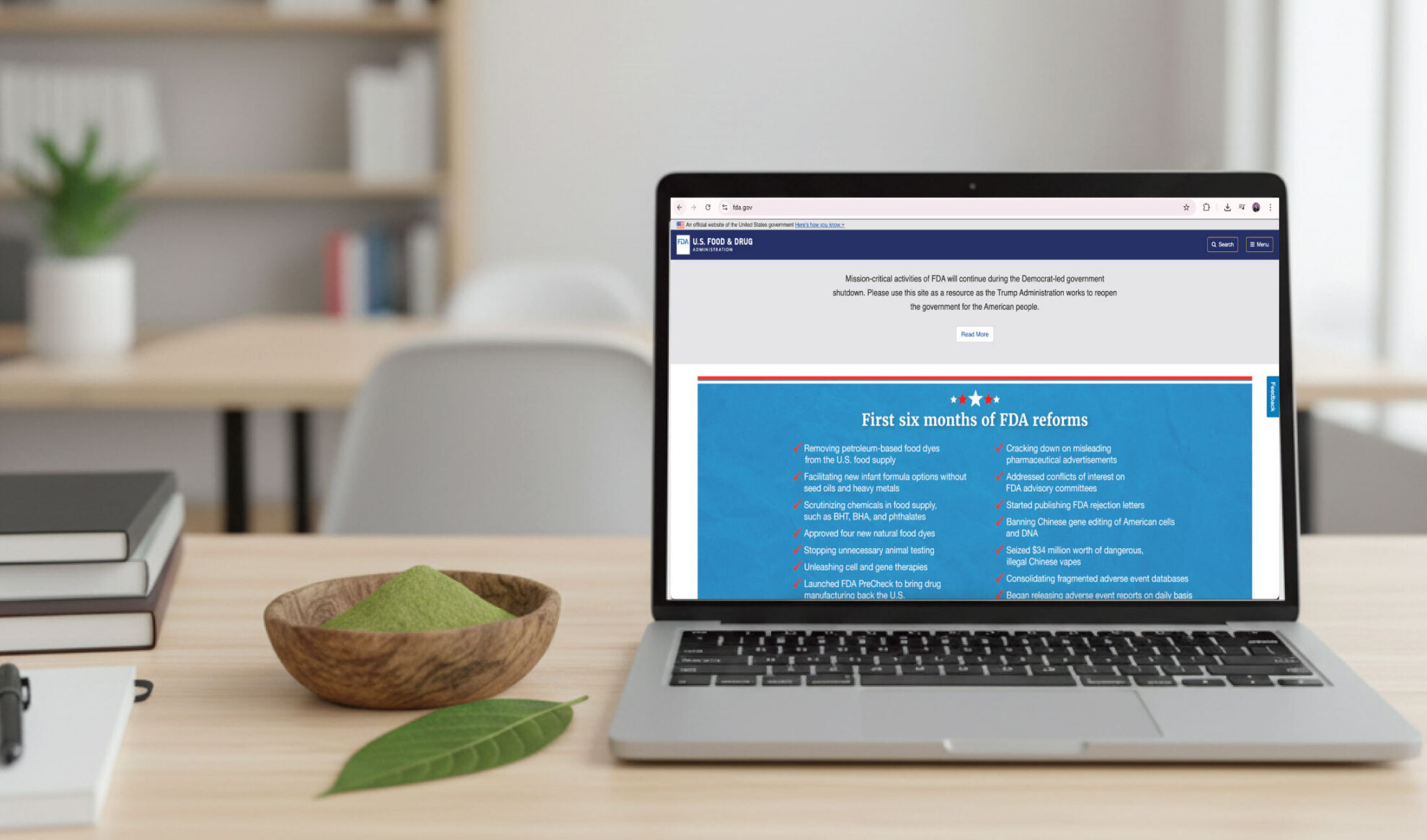 A laptop on a desk displays a website about FDA reforms. Next to the laptop are a bowl with green powder, a wooden tray, and a pen, with blurred plants and books in the background.