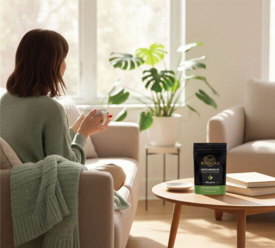 A woman sits on a sofa holding a mug, facing a bright room with plants. On a round coffee table in front of her is a black and green package labeled “Botanical’s White Maeng Da.”