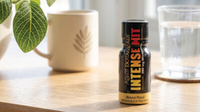 A small black bottle labeled Intense Mit sits on a wooden table near a potted plant, a mug, and a glass of water, with sunlight streaming through a window in the background.