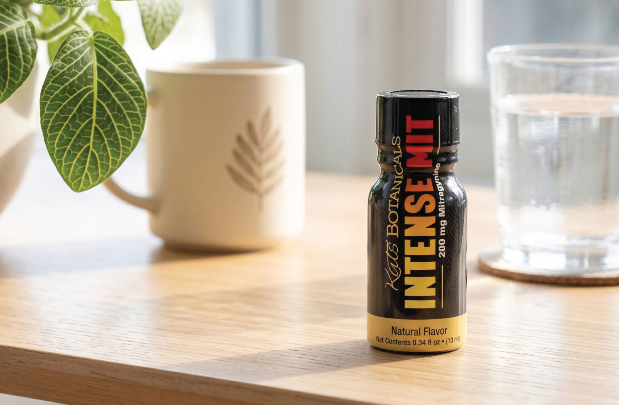 A small black bottle labeled Intense Mit sits on a wooden table near a potted plant, a mug, and a glass of water, with sunlight streaming through a window in the background.