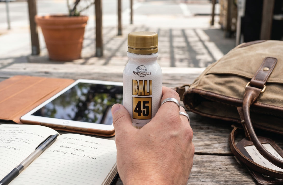 A hand holding a bottle labeled BALI 45 at an outdoor table with a tablet, notebook, pen, and tan bag nearby. Sunlight creates shadows across the scene.