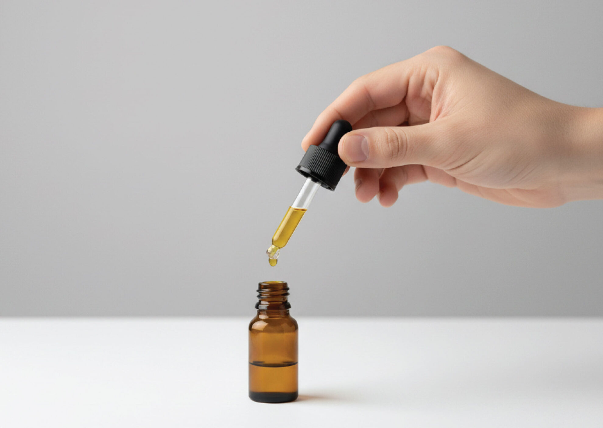 A hand holds a dropper above a small amber glass bottle, releasing a drop of yellow liquid, against a plain light gray background.