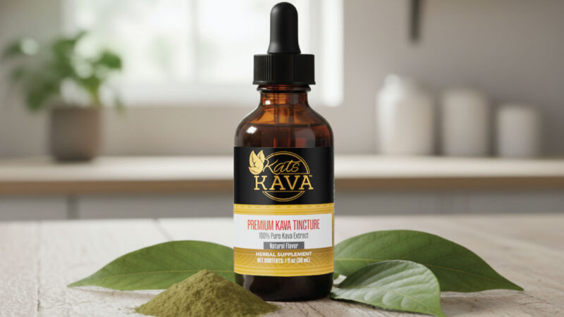 A brown glass dropper bottle labeled Kats Kava Premium Kava Tincture stands on a wooden surface, surrounded by kava leaves and a small pile of kava powder, with a blurred background.